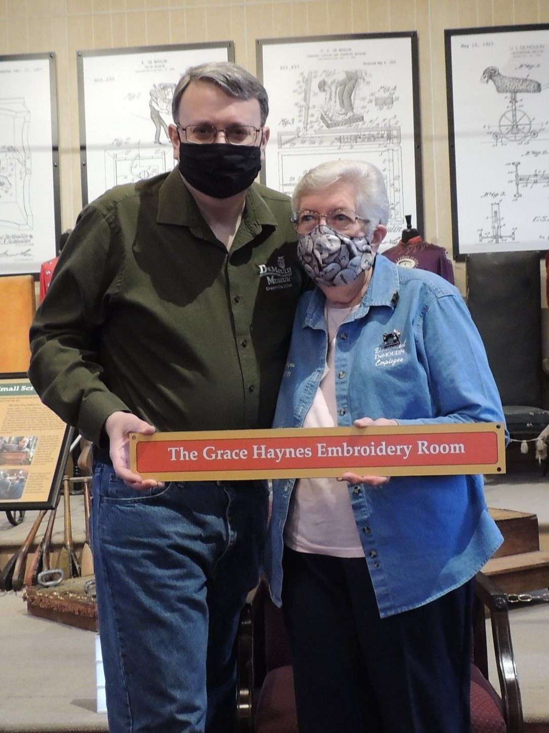 Man in green shirt and Woman in blue shirt stand together holding red and yellow museum sign.