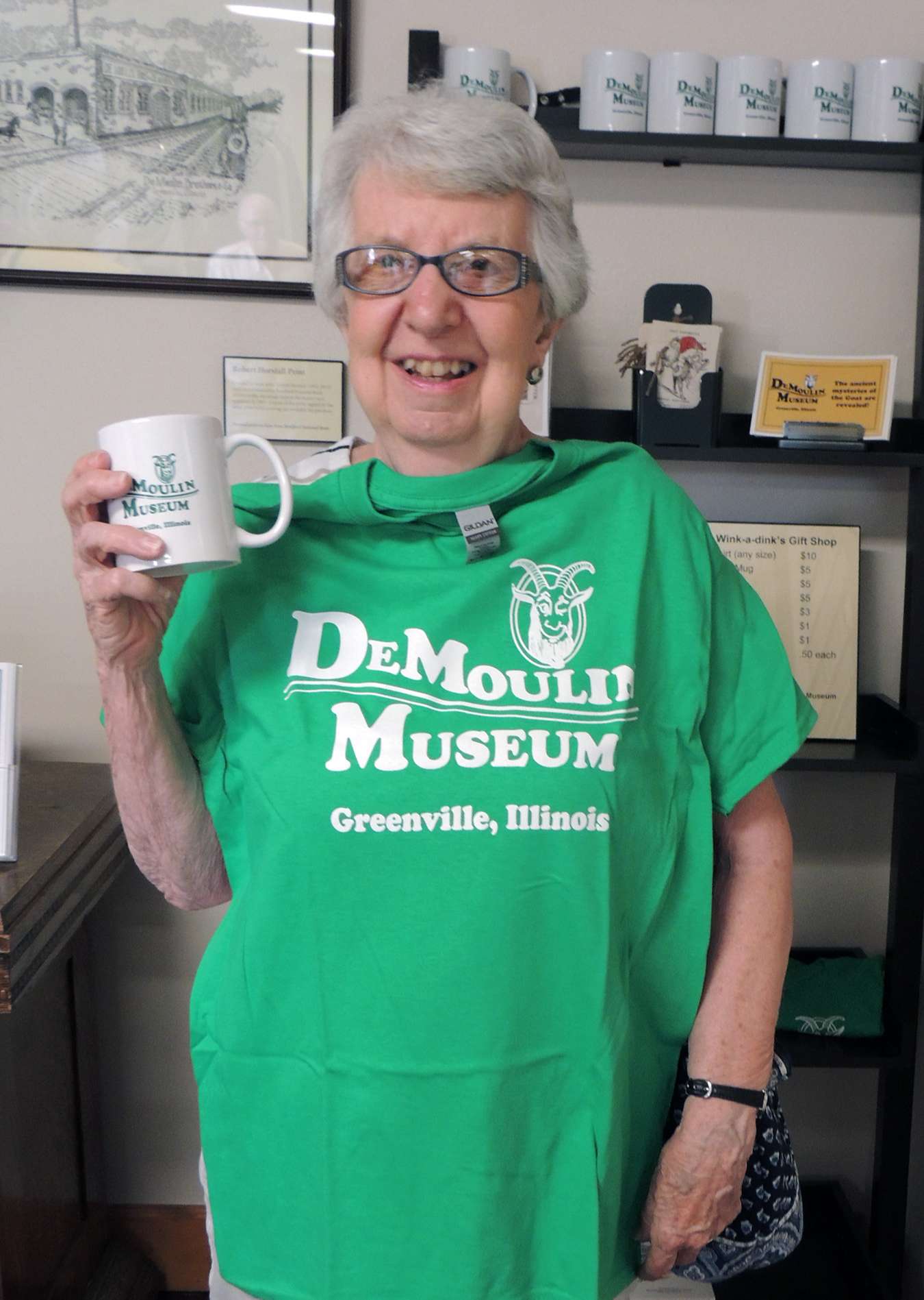 Smiling woman with white hair and eyeglasses holds white mug and green tee shirt