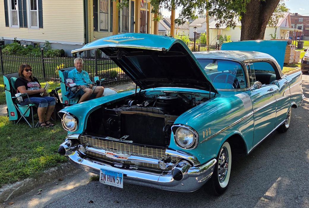 Vintage turquoise 1957 Chevrolet Bel Air with hood open, parked on a sunny street as two people admire its classic chrome details.