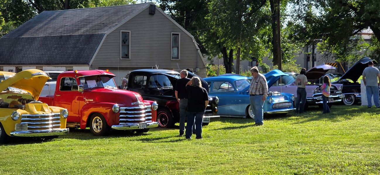 Outdoor vintage car show with colorful classic cars, visitors chatting on a sunny lawn, and a gray garage and trees in the background.