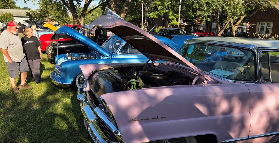 Classic car show on a sunny lawn, featuring a pink vintage Chevrolet with its hood open, surrounded by colorful classic cars and visitors.