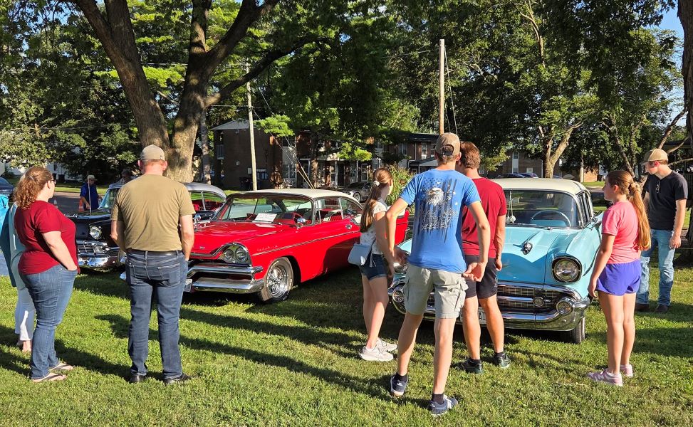 People gather at a sunny outdoor classic car show, admiring polished red and turquoise vintage cars in a leafy greenspace.