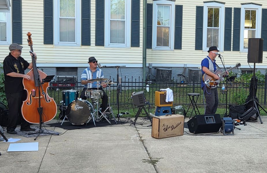 Three-piece jazz band performs outdoors: upright bassist, drummer, and guitarist sing beside microphones and speakers near a house.