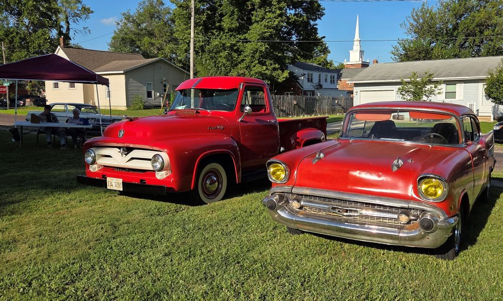 Bright red vintage pickup truck and classic sedan displayed on a sunny lawn, with trees, homes, picnic tables, and a church steeple behind.