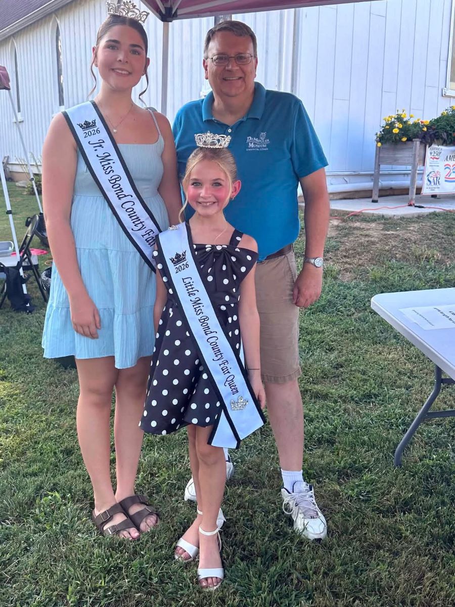 Smiling threesome poses outdoors: a teen pageant queen and young girl wear sashes and crowns beside a man at a community event.