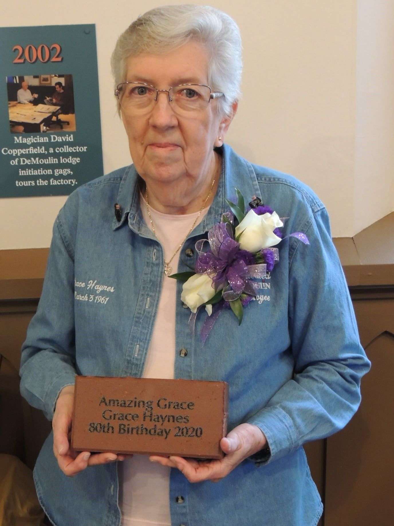 Woman with short gray hair and wire rimmed glasses holds red clay brick engraved with three lines of text