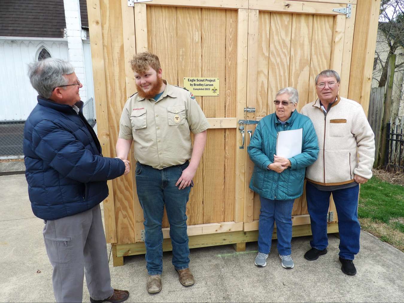 gray haired main in winter coat shakes hand of young man with red beard wearing tan scouting shirt in front of golden wood lumber shed door