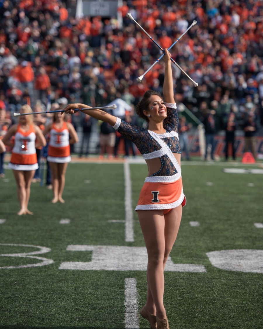 athletic young woman wearing orange and blue collegiate twirling uniform on green football field