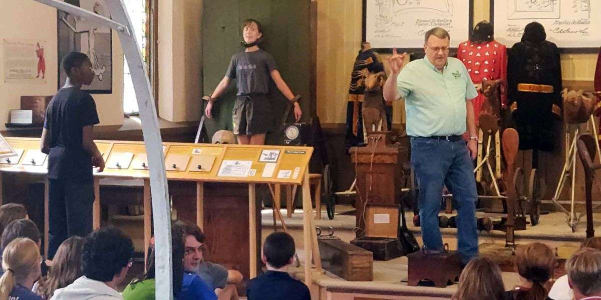 museum curator explains how the knife board works while a female high school student is strapped to it fearfully