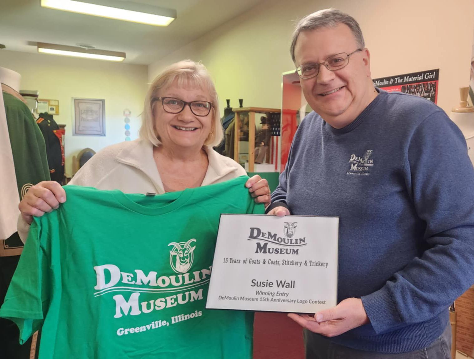 woman with bobbed blonde hair and eyeglasses holds a green museum tee shirt; and man with short gray hair and eyeglasses holds plaque