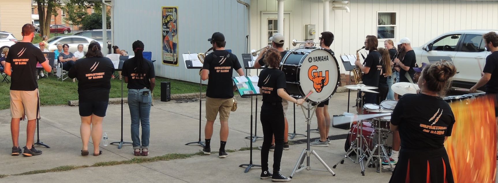 young men and women stand outside on concrete slab with musical instruments while older people listen to concert from lawn chairs.