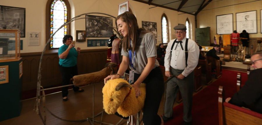 teenage girl grips yellow wool and brown horns of a model goat mounted on wheels as she's pushed through museum exhibit room, 2018