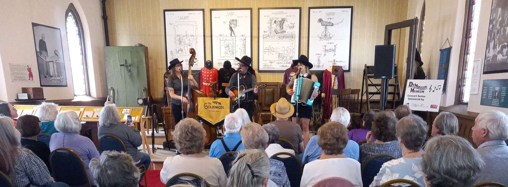 three musicians wearing black brimmed hats stand playing upright bass, guitar, and accordian in front of seated audience, sharing museum stage with lodge initiation devices like goats, trick stairs, trick chairs, and knife throwing board.
