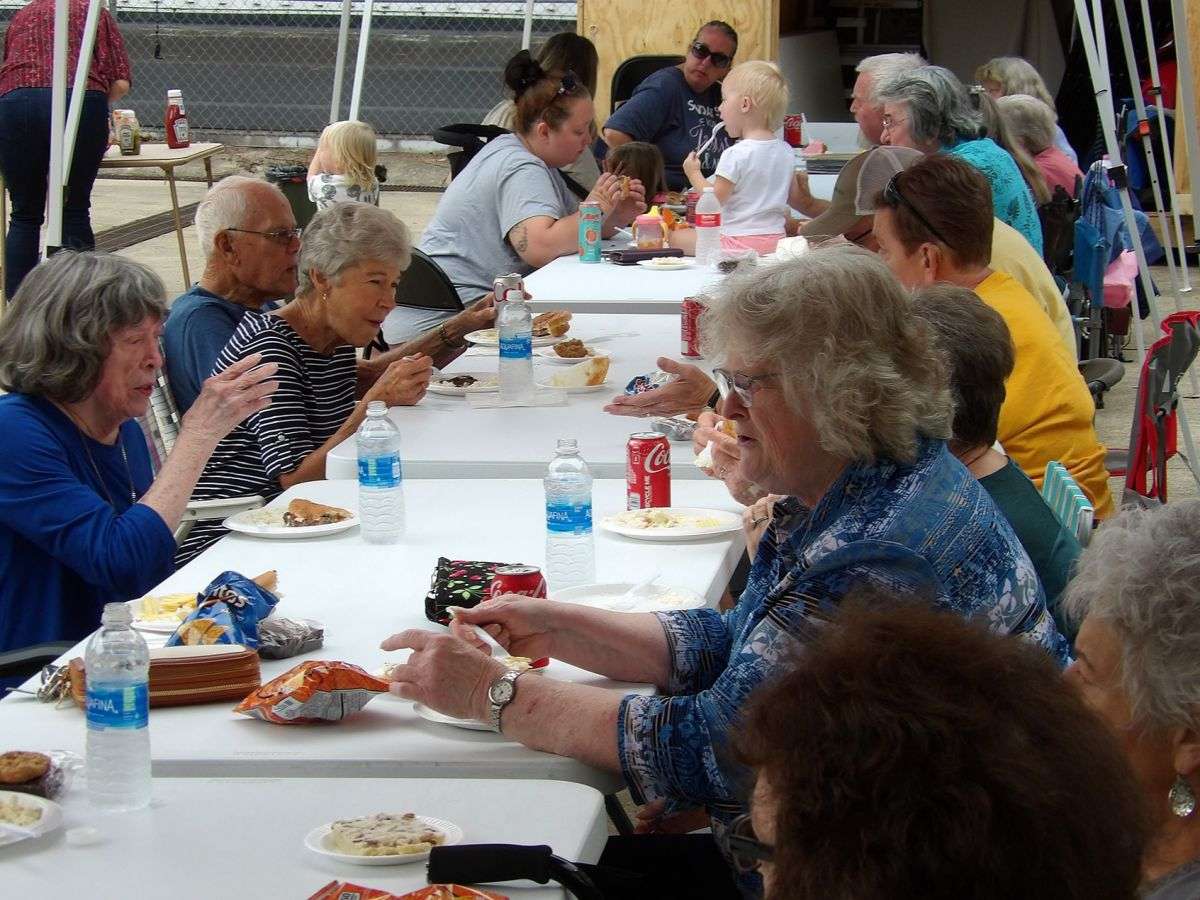 people of all ages eating lunch at long tables outside, renewing friendships and sharing memories.