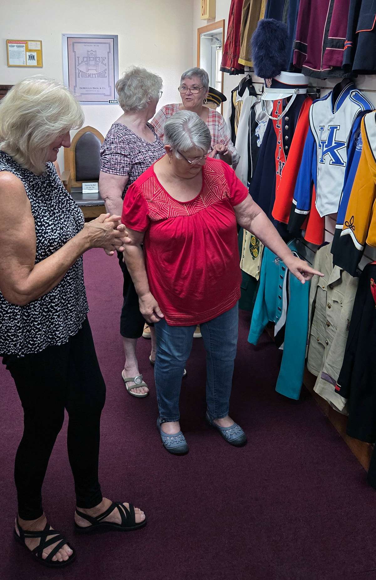 four women with gray hair converse and point to a wall display of hanging colorful band uniform jackets