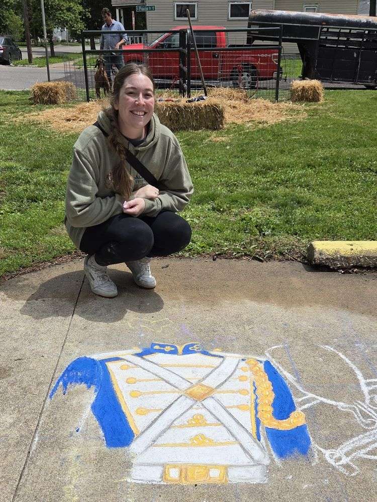 A woman kneels on a sidewalk next to a colorful chalk drawing of a blue and yellow marching band jacket. Hay bales and a red truck are in the background.