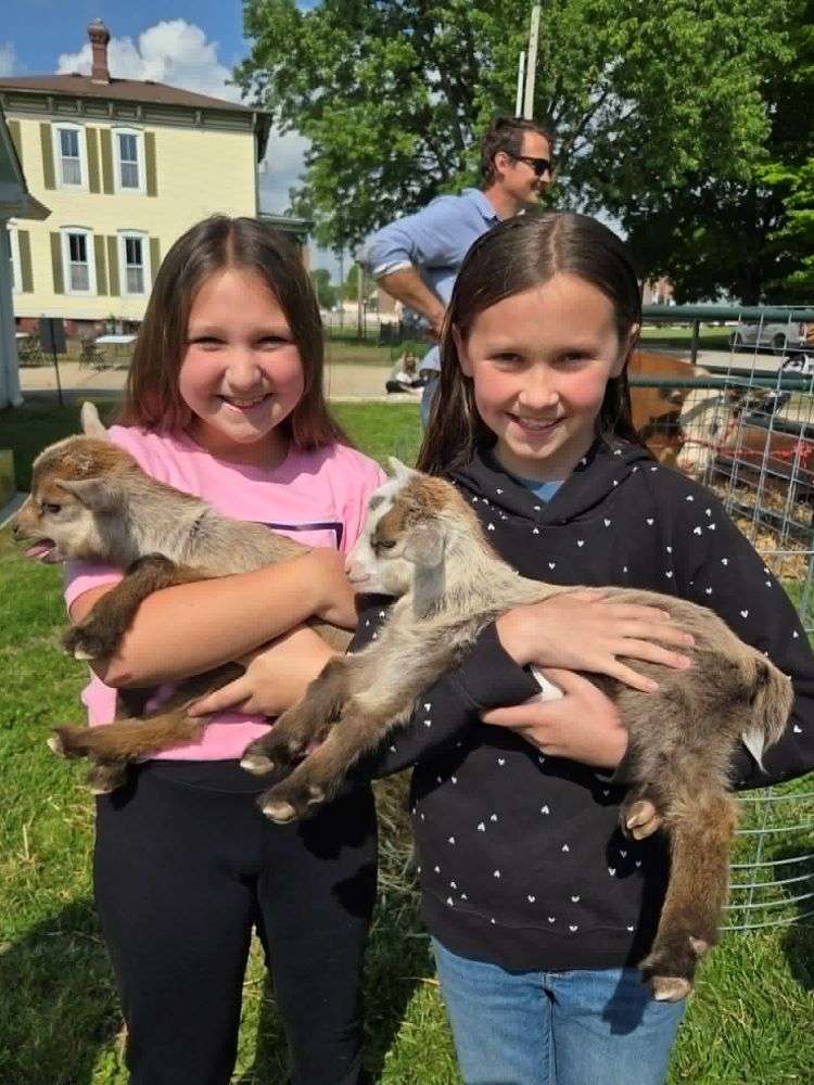 Two smiling girls hold baby goats, standing on grass. One wears a pink shirt, the other a dark polka-dot sweater. A sunny day, with a house and trees behind.