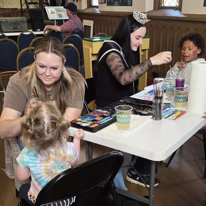 Two women paint children's faces at a table. One woman wears a tiara and sash, creating a playful atmosphere. The mood is joyful and creative.