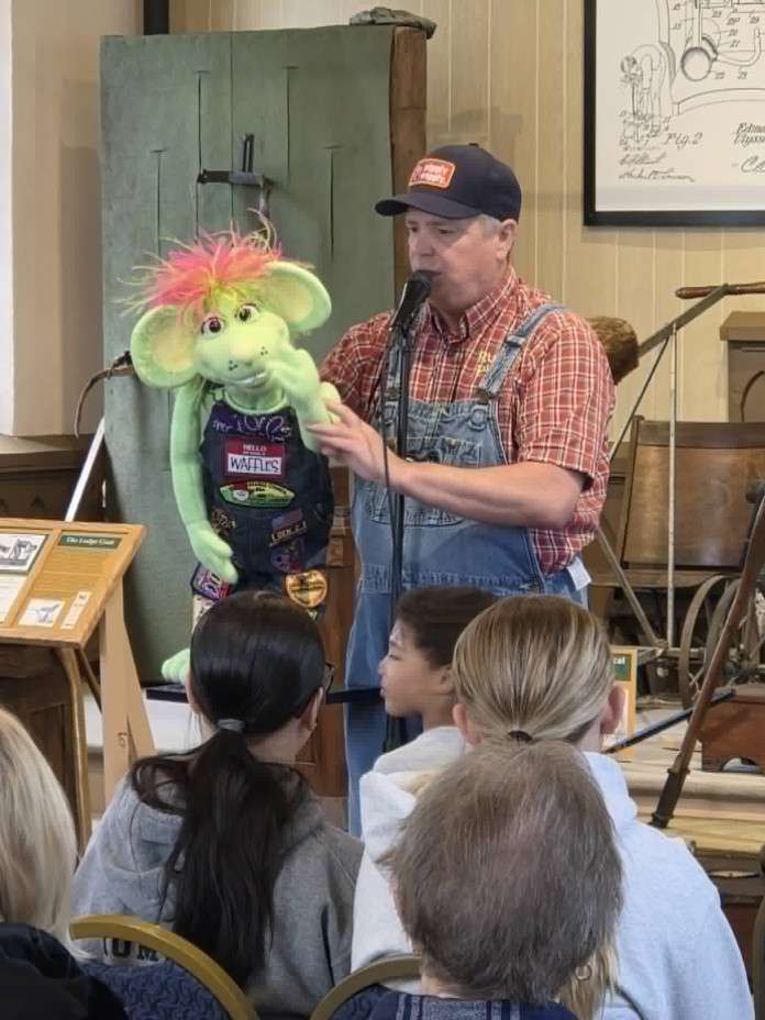 A man in overalls and a cap performs with a green puppet featuring colorful hair at a small indoor venue. Seated children watch attentively.