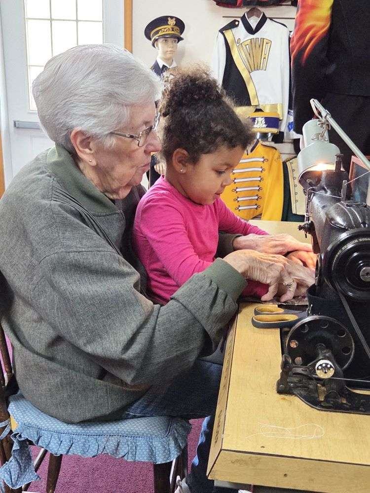 Elderly woman and young girl sewing together on an antique machine. Behind them, mannequins in marching band uniforms create a nostalgic atmosphere.