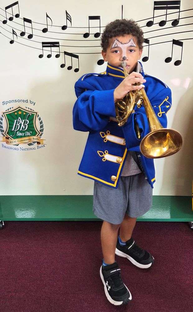 A young child in a blue marching band uniform with gold accents plays a trumpet. His face is painted with star designs, set against a musical notes backdrop.